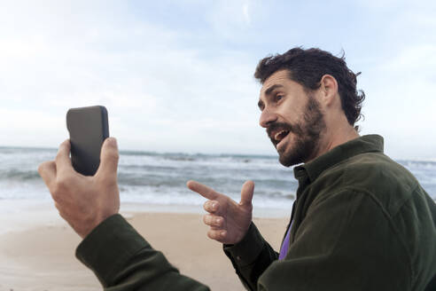 Mann gestikuliert und führt ein Videogespräch über ein Mobiltelefon am Strand - JOSEF23145