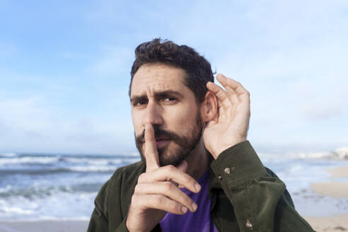 Man eavesdropping with finger on lips near sea at beach - JOSEF23151