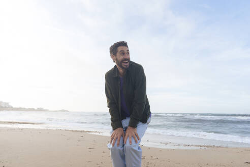 Fröhlicher Mann mit Händen auf Knien am Strand stehend - JOSEF23158