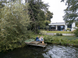 Family sitting on jetty with dog near trees - JOSEF23168