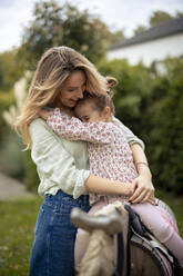 Mädchen sitzt auf Holzpferd und umarmt glückliche Mutter im Garten - JOSEF23171