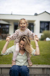 Happy mother holding hands with daughter sitting on shoulders in front of house - JOSEF23172