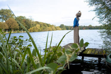 Frau steht auf einem Steg und wirft sich die Haare vor einem See zurecht - JOSEF23182