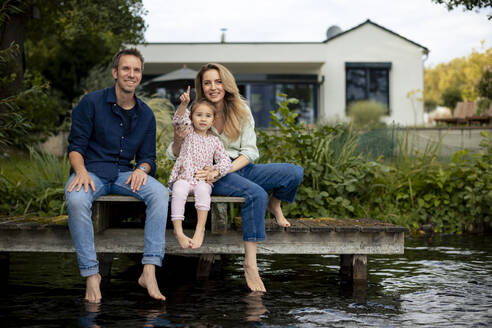 Smiling man and woman sitting near daughter gesturing near lake - JOSEF23208