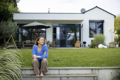 Smiling woman with eyes closed sitting in front of house - JOSEF23232