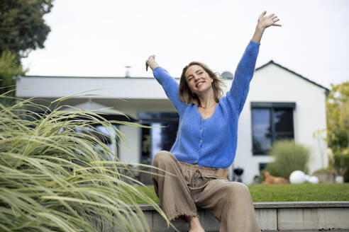 Carefree woman with arms raised in front of house - JOSEF23237