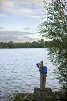 Frau mit Hand im Haar vor einem See auf einem Pier stehend - JOSEF23239