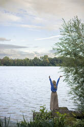 Carefree woman standing with arms raised near lake at sunset - JOSEF23241