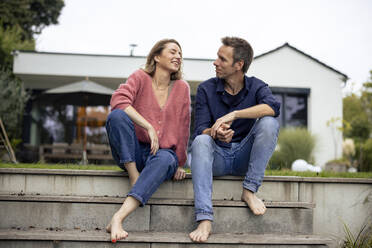 Happy couple sitting and talking to each other on steps in front of house - JOSEF23258