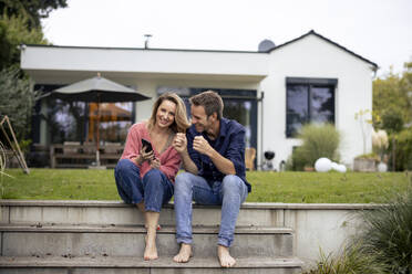 Happy couple sitting on steps with smart phone in front of house - JOSEF23259