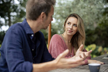 Man talking to woman sitting at table in garden - JOSEF23263