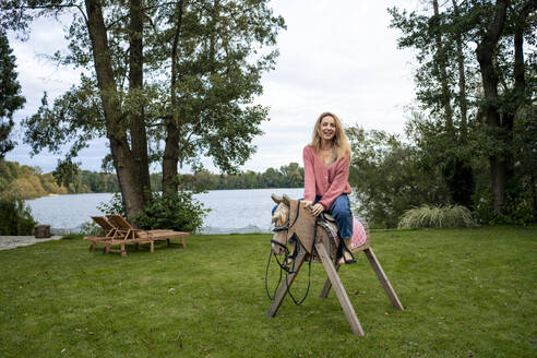 Glückliche Frau sitzt auf einem Holzpferd im Garten - JOSEF23284