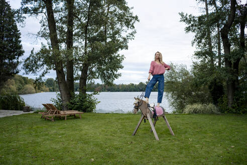 Woman standing on wooden horse in front of lake - JOSEF23286