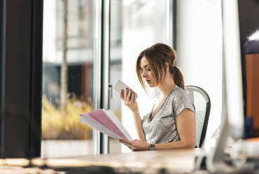 Geschäftsfrau prüft Dokumente und spricht mit einem Smartphone im Büro - UUF31113