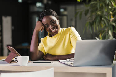 Lächelnde Geschäftsfrau, die sich im Büro auf einen Ellbogen neben einem Laptop stützt - UUF31134