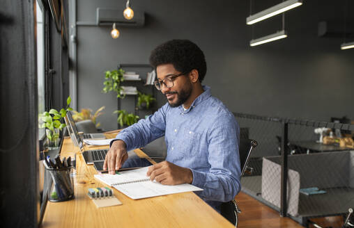 Businessman examining notes in diary near laptop at desk - VRAF00297