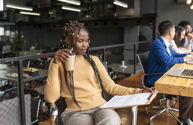 Businesswoman sitting with clipboard and coffee cup near colleagues in office - VRAF00314