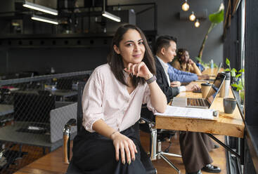 Smiling businesswoman with hand on chin sitting near colleagues at desk - VRAF00325