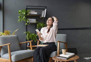 Happy businesswoman sitting with smart phone on armchair in office - VRAF00329