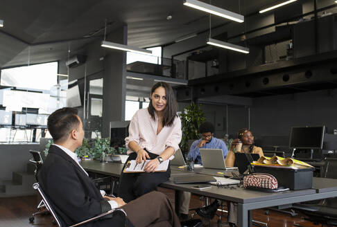 Lächelnde Geschäftsfrau, die auf dem Schreibtisch sitzt und sich mit einem Kollegen im Büro unterhält - VRAF00355