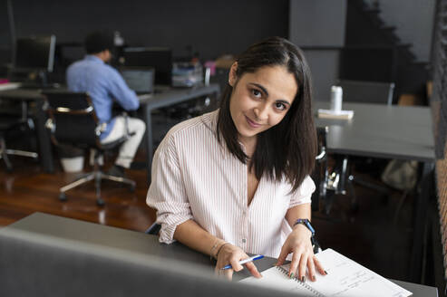 Smiling businesswoman sitting with diary at desk - VRAF00374