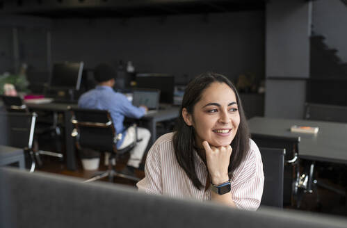 Smiling businesswoman sitting with hand on chin in office - VRAF00375