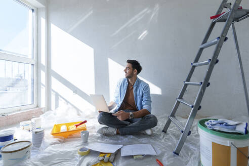 Junger Mann sitzt mit Laptop auf dem Boden in einem renovierten Zimmer - AAZF01443