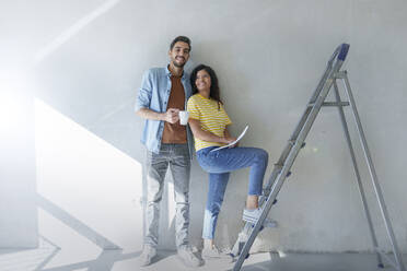 Happy couple standing in front of wall near ladder at new home - AAZF01455