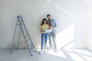 Young couple with tablet PC discussing over blueprint in front of wall - AAZF01459