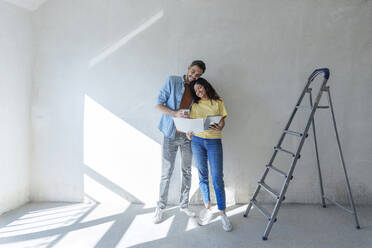 Happy couple with tablet PC analyzing blueprint near ladder at new home - AAZF01460