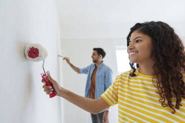 Lächelnde Frau, die mit einem Roller in der Nähe eines Mannes im neuen Haus eine Wand streicht - AAZF01466