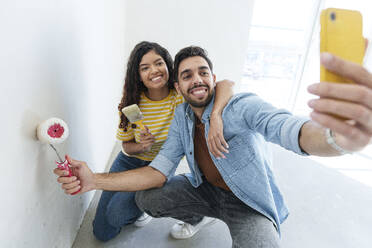 Verspieltes Paar mit Farbrolle und Pinsel, das ein Selfie mit seinem Smartphone in seinem neuen Haus macht - AAZF01470