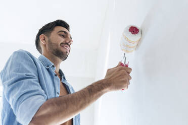 Lächelnder junger Mann, der eine Wand mit einem Farbroller streicht - AAZF01475