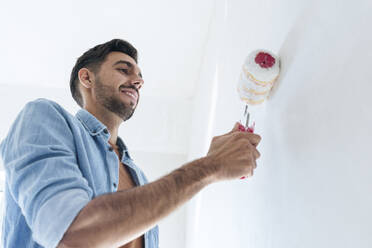 Smiling young man painting wall with paint roller - AAZF01475