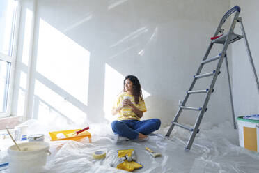 Smiling young woman with coffee cup sitting in room under renovation - AAZF01478