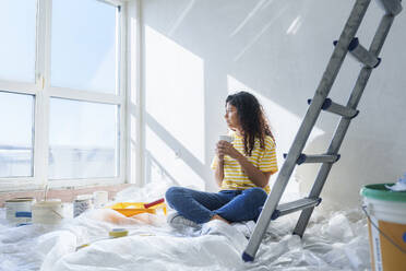 Young woman with coffee cup sitting near ladder at new home - AAZF01479