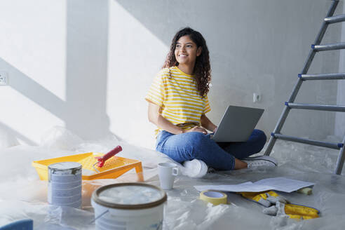 Smiling woman sitting on floor with laptop in room under renovation - AAZF01484