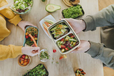 Hands of man holding vegetarian lunch box near woman at table - YTF01687