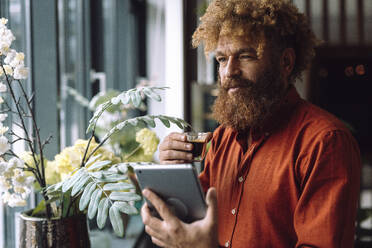 Businessman holding tablet PC and cup of black coffee near plants in office - JOSEF23308