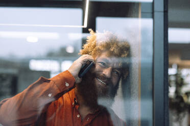 Happy mature businessman talking on mobile phone seen through glass - JOSEF23321
