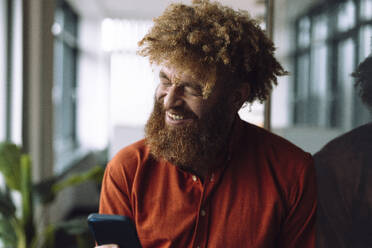 Happy businessman with smart phone near glass wall at office - JOSEF23324