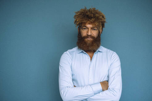 Mature businessman with arms crossed standing in front of blue wall - JOSEF23338