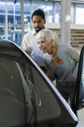 Verkäufer zeigt einer älteren Frau im Ausstellungsraum ein Auto - IKF01587