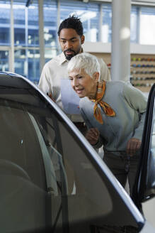 Verkäufer zeigt einer älteren Frau im Ausstellungsraum ein Auto - IKF01587
