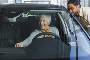 Lächelnde Verkäuferin mit Frau, die ein Auto im Ausstellungsraum begutachtet - IKF01599