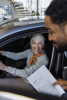 Lächelnde Verkäuferin hält Dokument in der Nähe des im Auto sitzenden Kunden - IKF01605