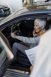 Smiling senior woman talking on smart phone in car at showroom - IKF01606