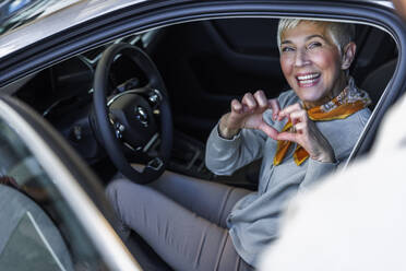 Lächelnde Frau gestikuliert in Herzform und sitzt im Auto im Ausstellungsraum - IKF01611