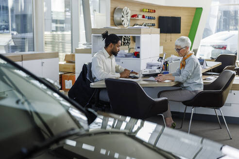 Kunde erledigt Papierkram mit Verkäufer im Autohaus sitzend - IKF01622