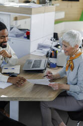 Happy senior woman discussing with salesperson over document at desk - IKF01633